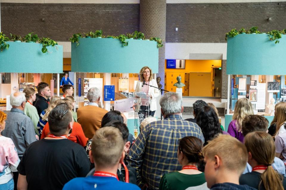 Kamervoorzitter Vera Bergkamp, achter haar de drie vitrines van de expositie en voor haar een groep bezoekers