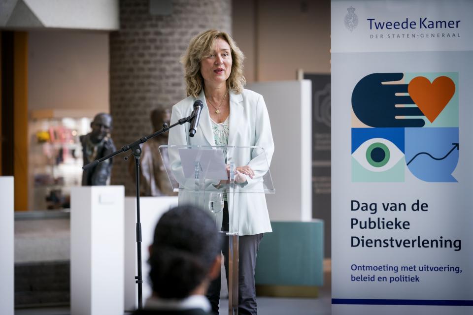 Kamervoorzitter Vera Bergkamp spreekt voor een microfoon in de Statenpassage. Achter haar staat een banner van de Dag van de Publieke Dienstverlening