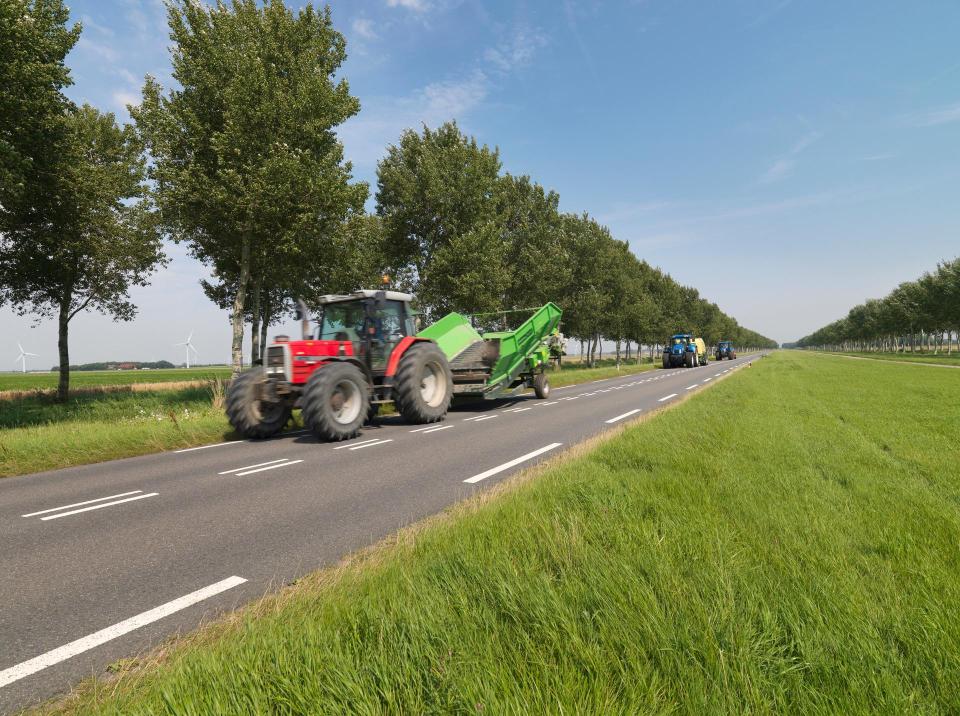 Tractor rijdt op provinciale weg, op de voorgrond een grasberm en achter de tractor bomen.