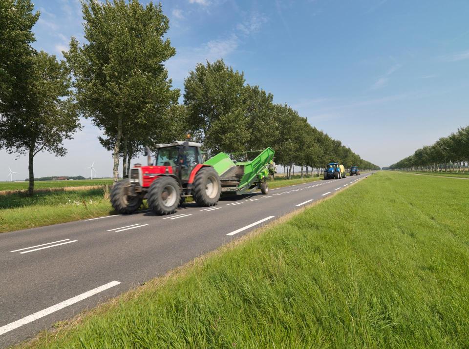 Tractor rijdt op provinciale weg, op de voorgrond een grasberm en achter de tractor bomen. 