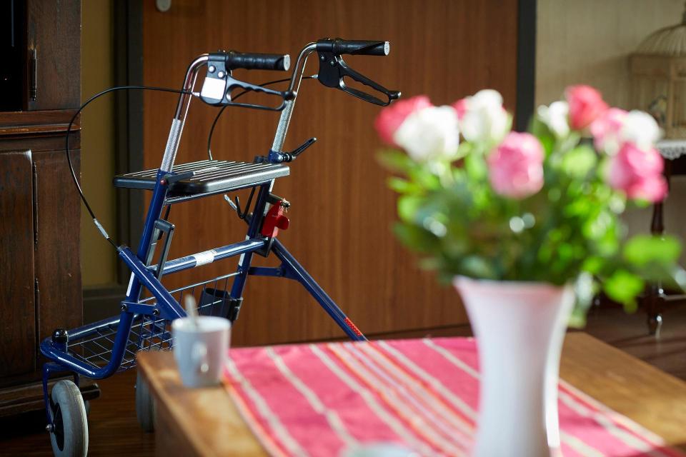 Op de voorgrond een tafel met daarop een vaas bloemen en een kopje, erachter een rollator.