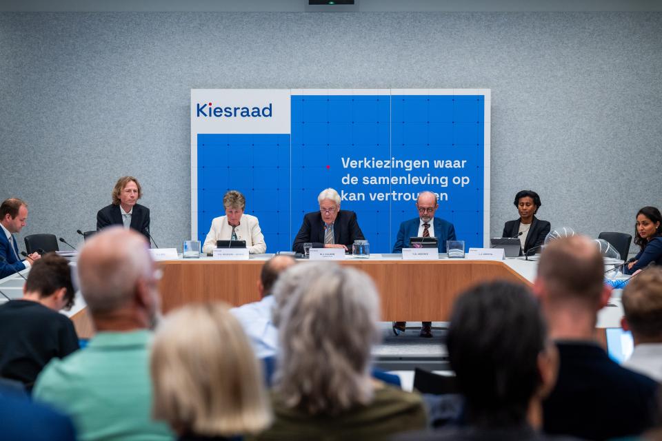 Beeld uit een zaal, publiek op de rug gezien. Achter een tafel voorin de ruimte zitten de vertegenwoordigers van de Kiesraad. Achter hen hangt een bord met de tekst: Verkiezingen waar de samenleving op kan vertrouwen.