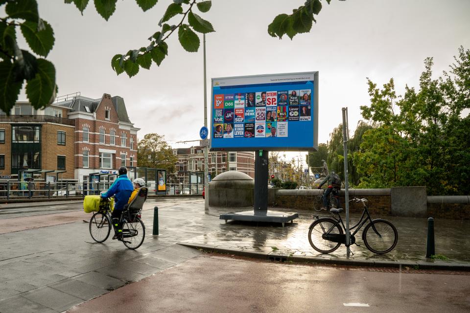 Regenachtig stadsbeeld met in het midden een groot bord met verkiezingsposters. Links ervoor een fietser met een kind achterop. 