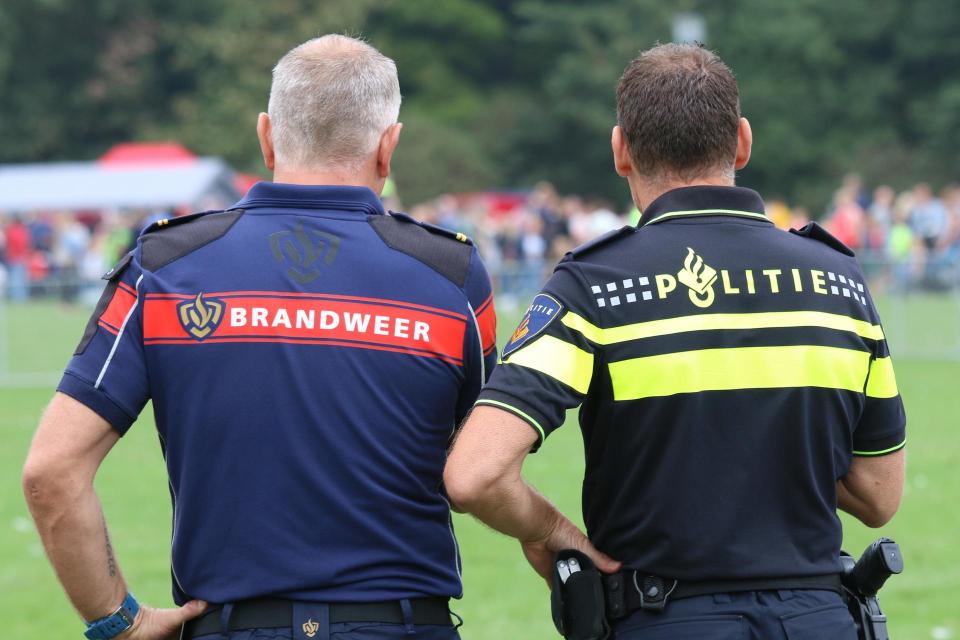 Een brandweerman en een politieman naast elkaar op de rug gezien. Achter op hun shirts staat: Brandweer en Politie.