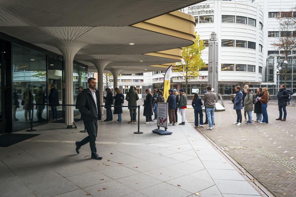 Beeld vanaf de zijkant van de publieksingang Tweede Kamer. Een rij van zo'n twintig wachtenden op de achtergrond en iemand die het pand verlaat op de voorgrond. 