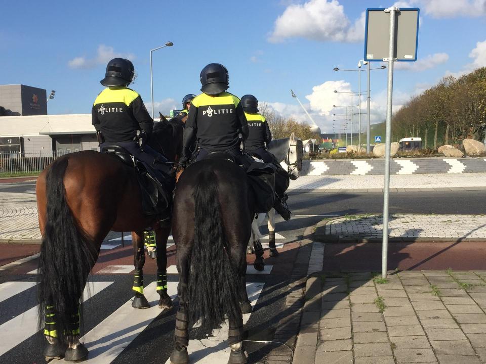 Drie paarden met politieagenten, van achteren gezien, lopen in de richting van een voetbalstadion.