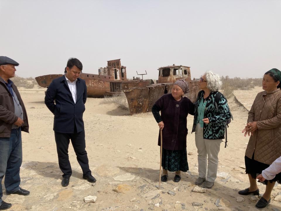 Eerste Kamerlid Karimi is in gesprek met verschillende personen op een zandvlakte in Muynak. Er zijn scheepswrakken op de achtergrond te zien.