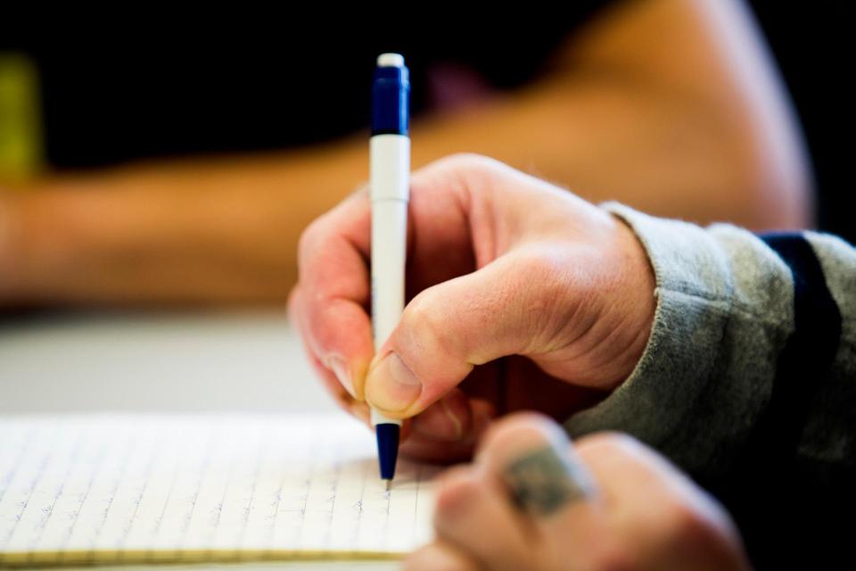 De rechterhand met pen schrijft op een notitieblok, de linkerhand rust naast het blok op tafel. Hierachter is de onderarm van iemand anders te zien. 