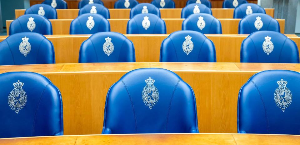 Overzicht van vier rijen blauwe zetels achter de Kamerbankjes in de plenaire zaal van de Tweede Kamer. 