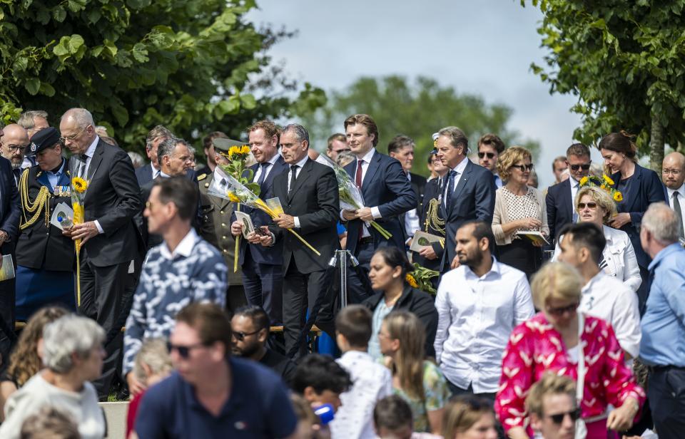 Een rij officiële vertegenwoordigers van de overheid en uit het buitenland loopt met ieder een zonnebloem in de hand in een rij achter elkaar. Op de voorgrond andere aanwezigen. 