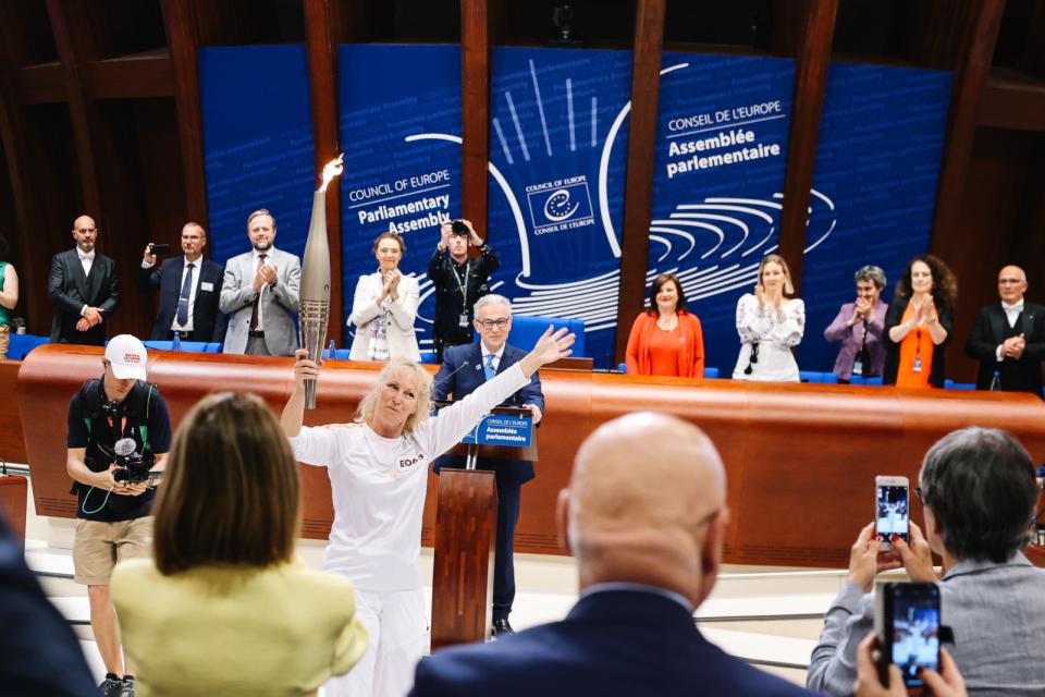 De olympische vlam deed op 26 juni de plenaire vergaderzaal aan. Op de foto is te zien hoe een mevrouw de fakkel in haar hand heeft. Ze wordt omringd door mensen die applaudiseren.