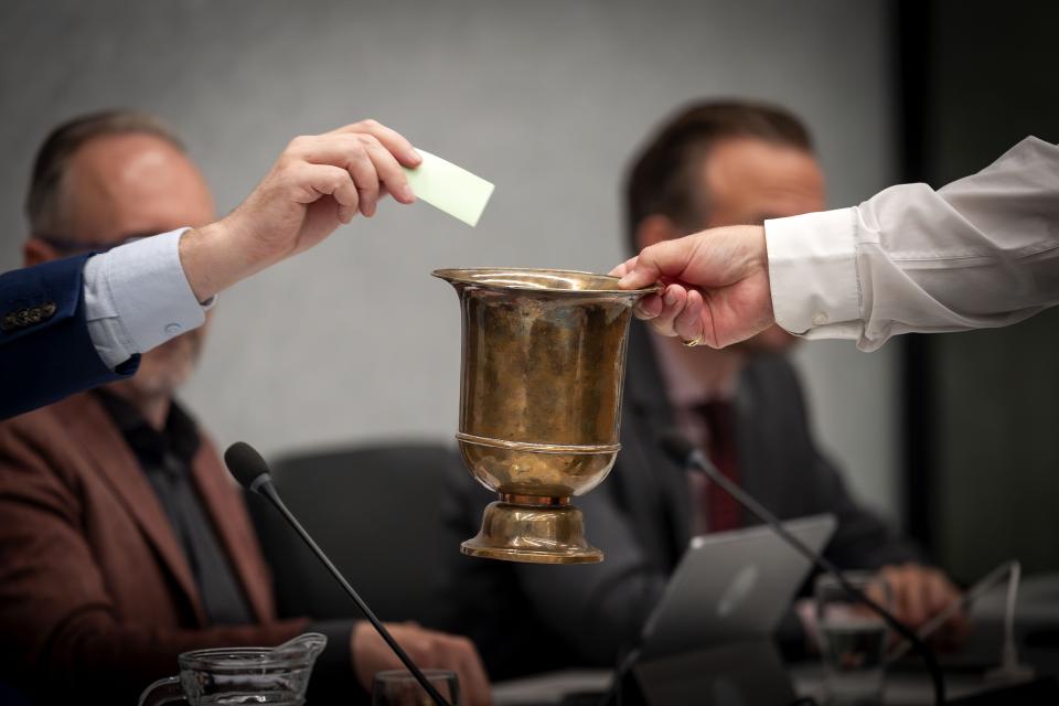 Op de voorgrond een uitgestoken hand met een dichtgevouwen papier boven een beker. Op de achtergrond kijken Kamerleden toe.