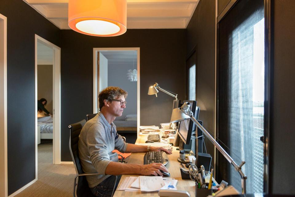 Man zit aan computer op overloop, achter hem is een slaapkamer met bedden zichtbaar.