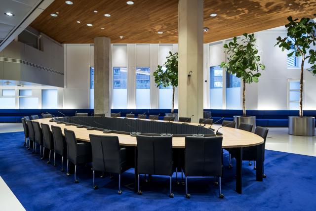 Grote ovale vergadertafel in het midden van een zaal op een hardblauw vloerkleed met ongeveer twintig stoelen er omheen.
