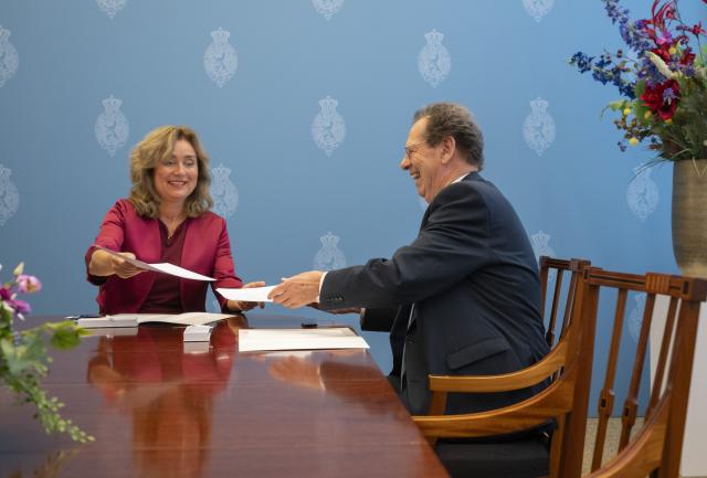 Kamervoorzitter Vera Bergkamp zit links aan een tafel in de Schrijfkamer en verkenner Gom van Strien rechts. Ze wisselen documenten uit. 