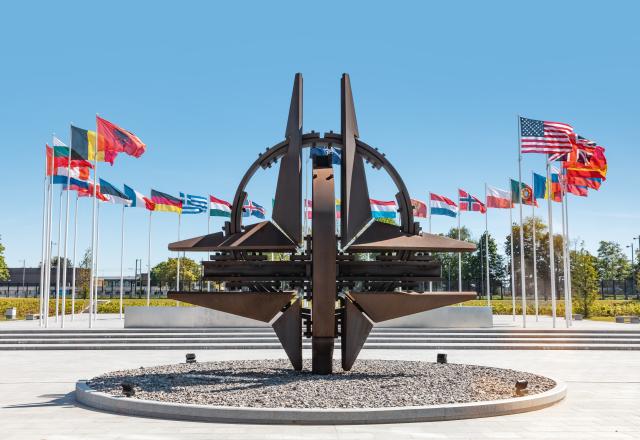 Op de voorgrond staat een monument dat bestaat uit het NAVO-symbool. Op de achtergrond wapperen een aantal vlaggen aan vlaggenmasten. 