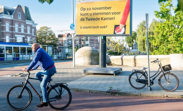 Stadsbeeld: een man fietst langs een bord waarop staat: Op 22 november zijn er verkiezingen voor de Tweede Kamer. U komt toch ook?