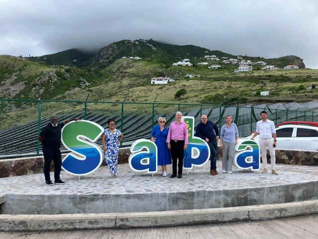 Voor een groene heuvel staan op een plateau langs de snelweg vier kunststof letters die samen het woord Saba vormen. De commissieleden, gezaghebber en eilandsecretaris poseren tussen de letters. 