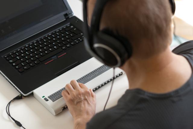 Een persoon die werkt op een laptop met brailleleesregel en koptelefoon.